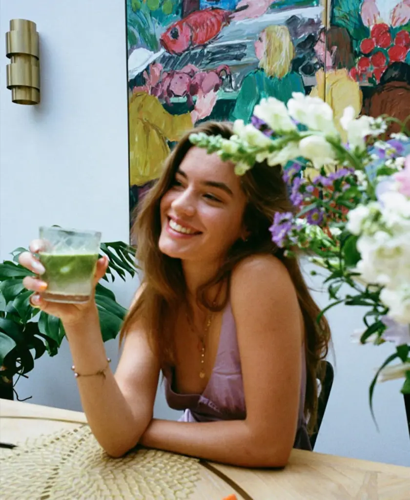 Smiling woman holding a glass of green drink beside a colorful flower arrangement