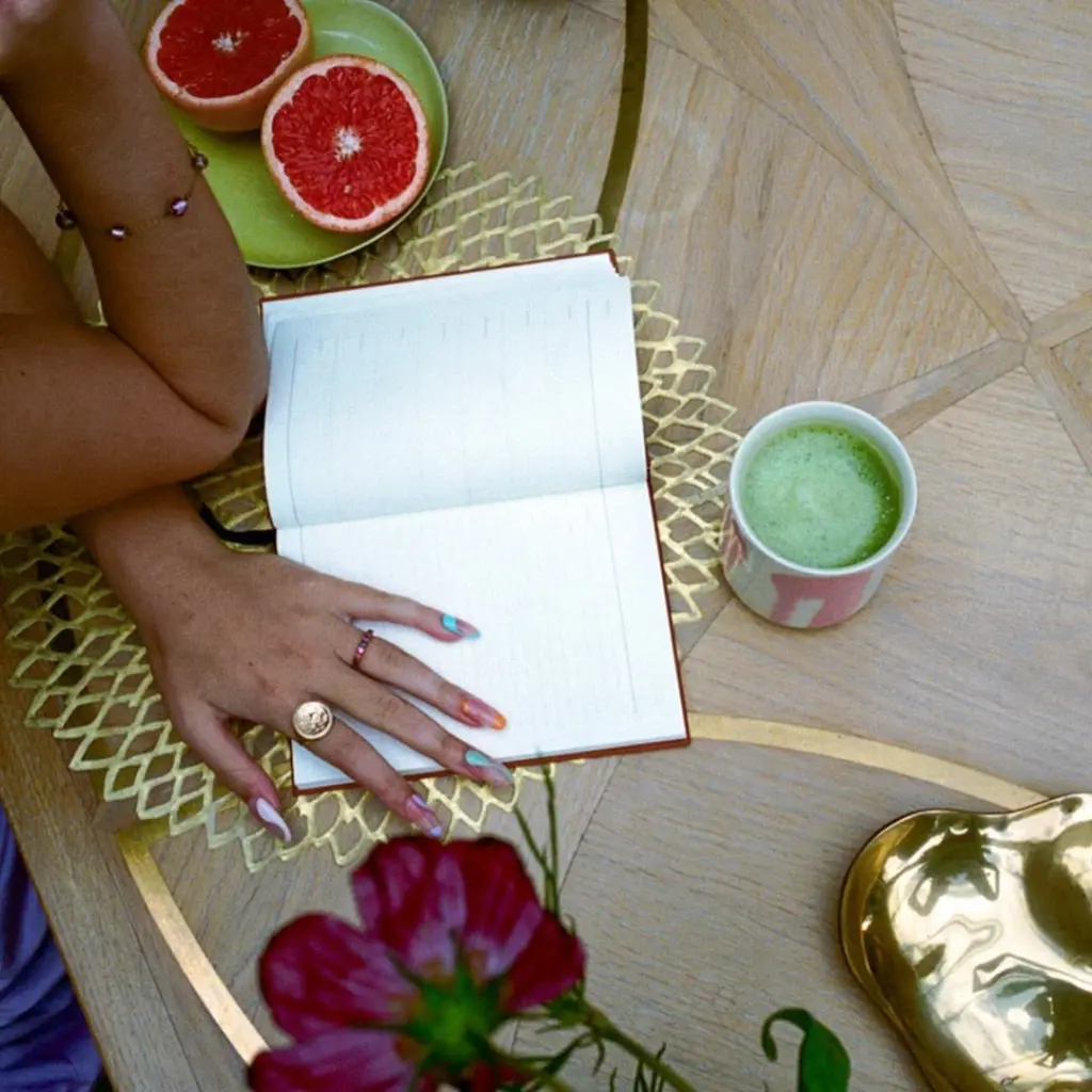 Hands beside an open notebook, a cup of green drink, and halved grapefruit