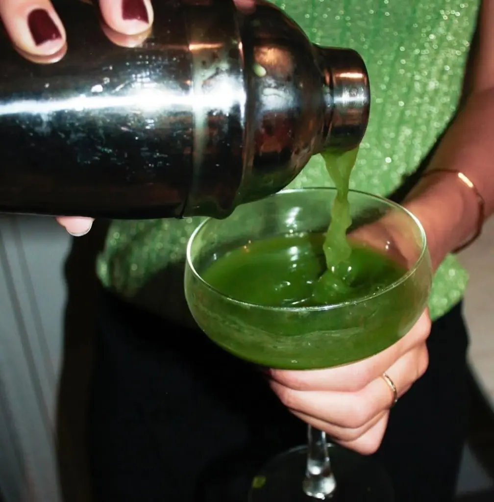 Green drink being poured from a metal shaker into a coupe glass