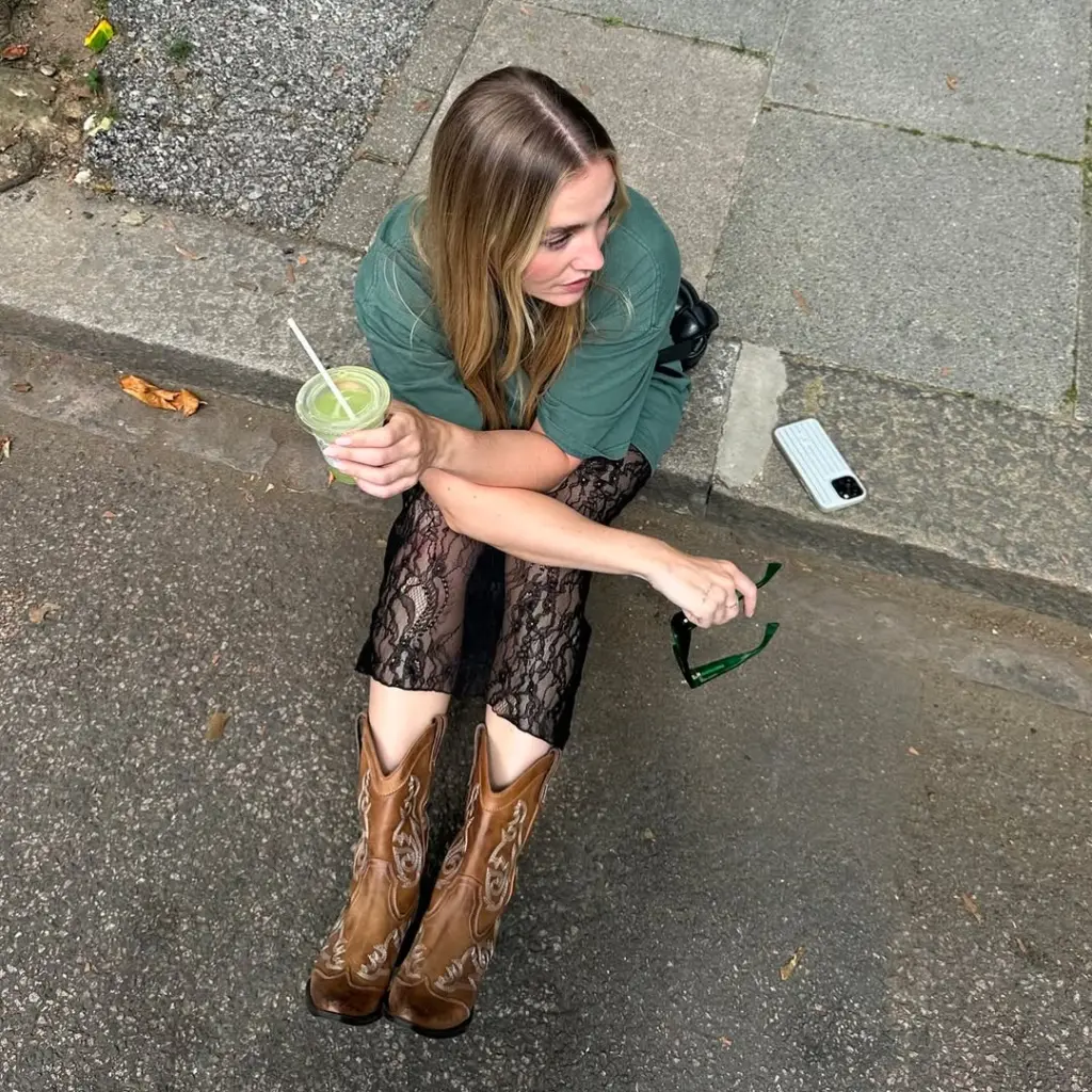 Woman in brown cowboy boots sitting on a curb with a green drink and sunglasses