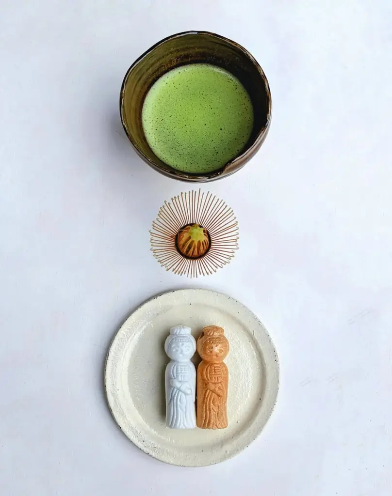 Green matcha in a ceramic bowl with a bamboo whisk and two figurine-shaped sweets