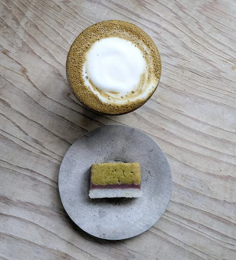 Foamy brown tea beside a small layered sweet on a gray plate