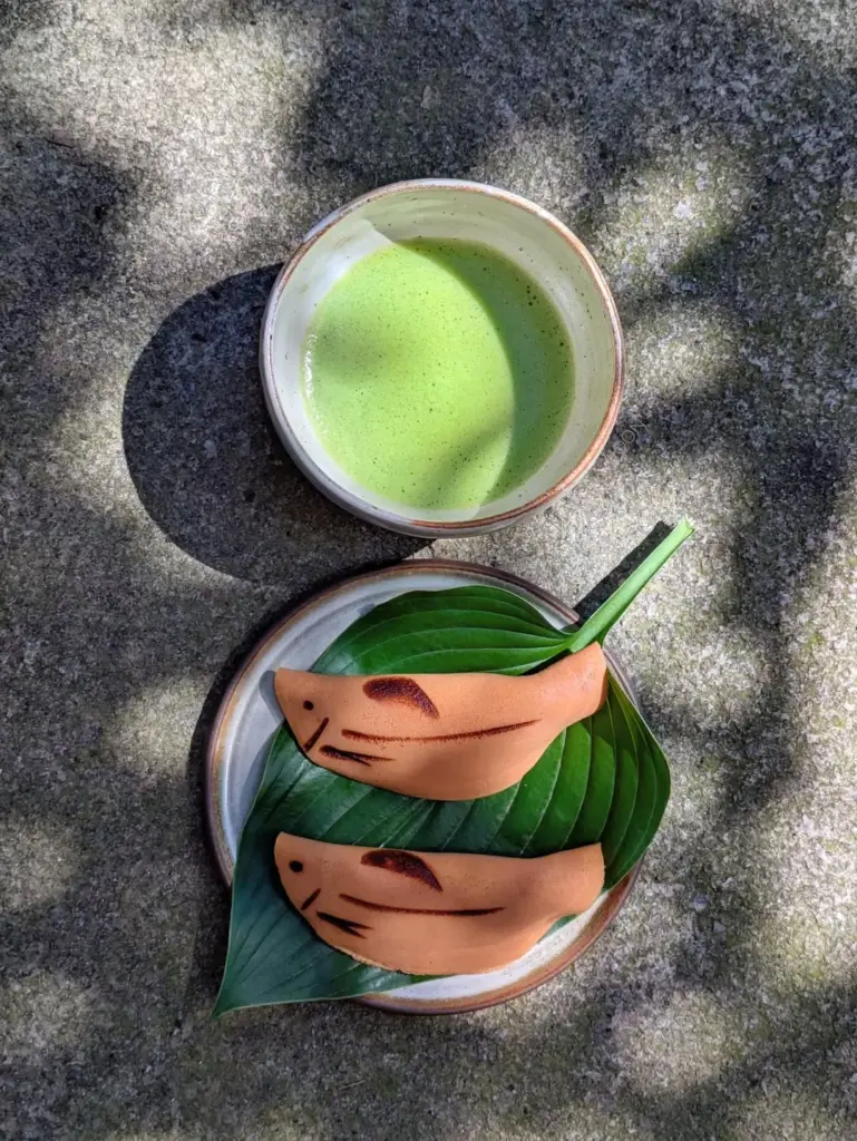 Bowl of bright green matcha beside two fish-shaped sweets on leaves
