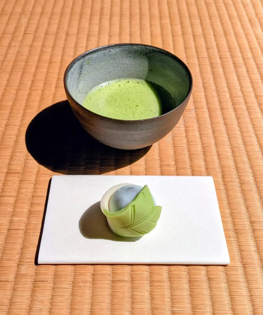 Ceramic bowl of frothy green tea beside a green leaf-shaped sweet on tatami
