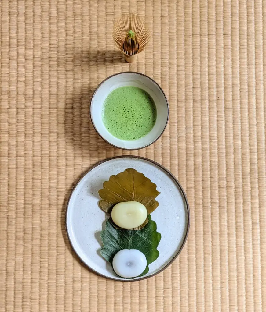 Bowl of frothy green tea with two leaf-wrapped sweets and a bamboo whisk