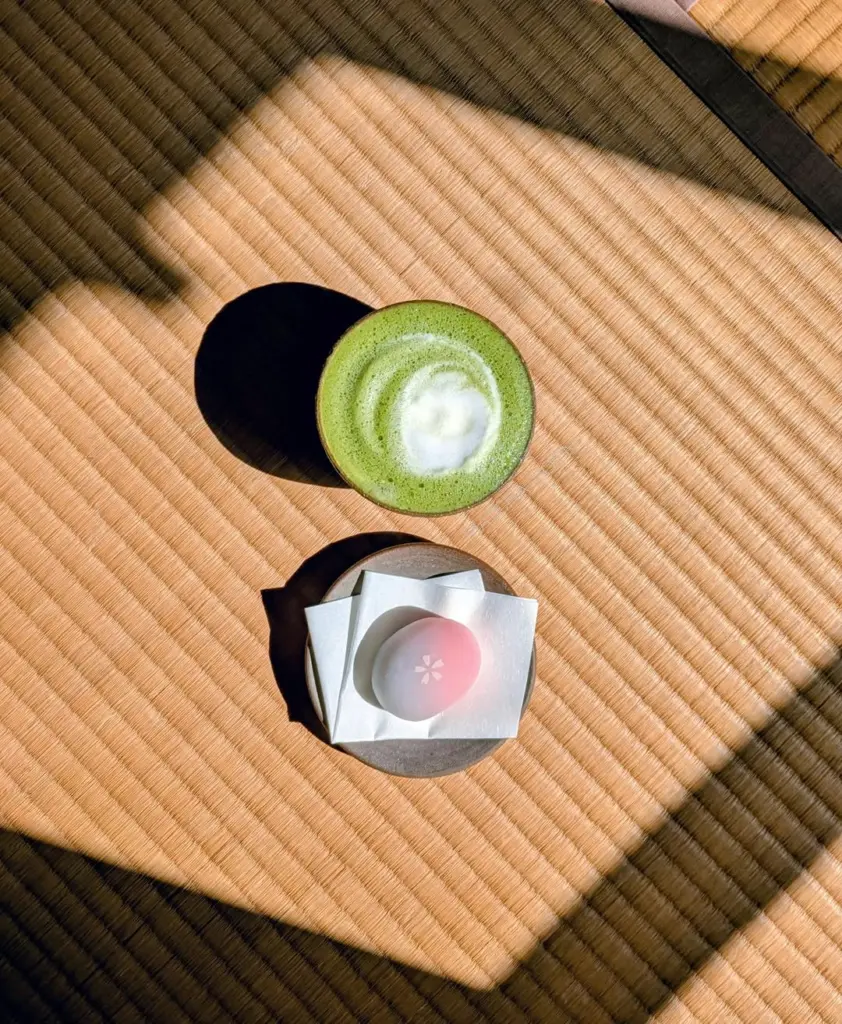 Bowl of frothy green tea and a pink flower-shaped sweet on a sunlit tatami mat