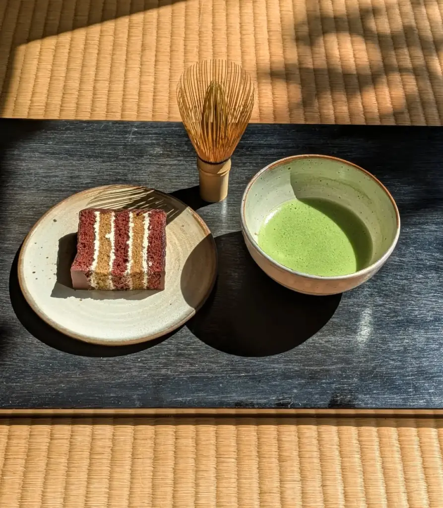 Green matcha in a ceramic bowl beside a bamboo whisk and slice of layer cake