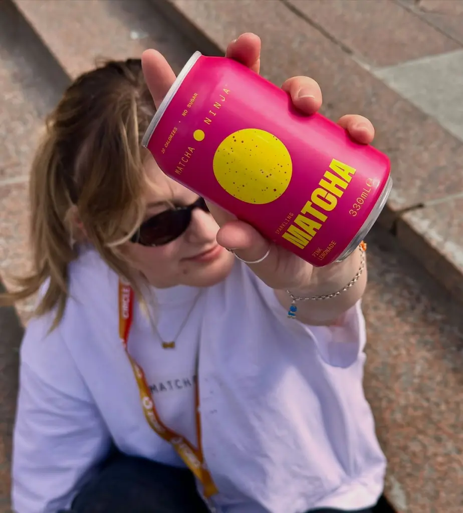 Person in sunglasses holding a bright pink Matcha Ninja can toward the camera