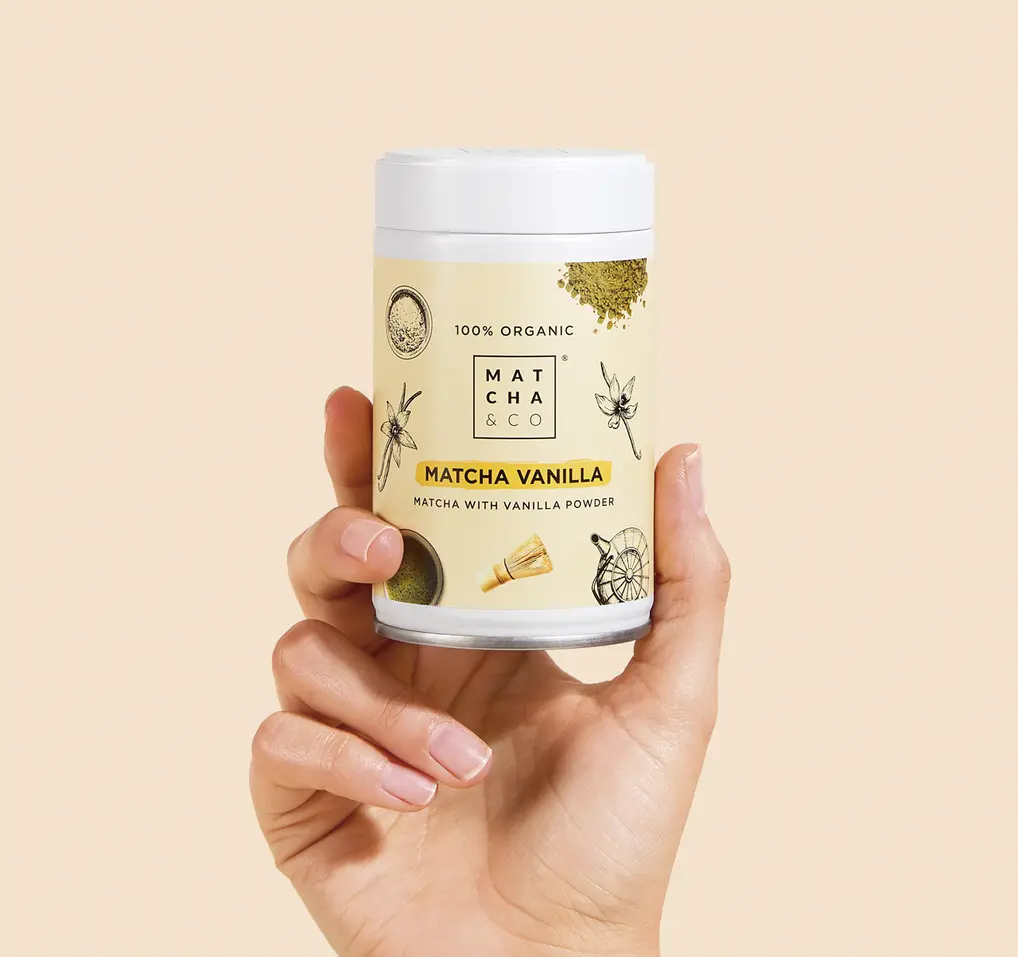 Hand holding a Matcha & CO Matcha Vanilla container against a pale beige background