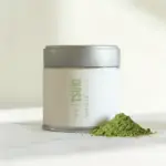 Silver-lidded tin with cream label reading Tsuki Matcha Vanille beside a mound of green matcha powder on a marble surface