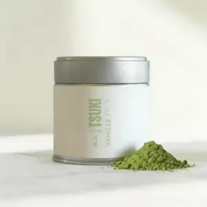 Silver-lidded tin with cream label reading Tsuki Matcha Vanille beside a mound of green matcha powder on a marble surface