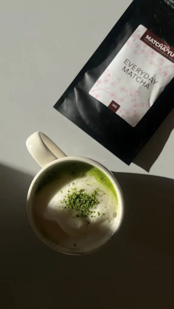 Foamy matcha latte topped with green powder beside an Everyday Matcha pouch