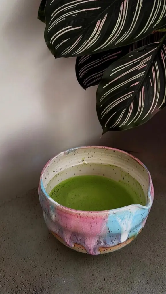 Green matcha in a pink and blue glazed ceramic bowl beneath striped leaves