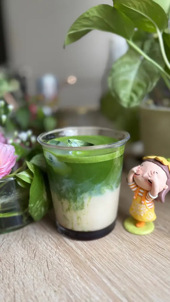 Layered iced matcha latte in a clear glass beside leafy plants and a small figurine