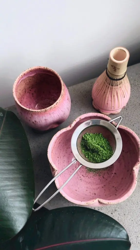Green matcha powder in a sieve over a pink bowl beside a bamboo whisk