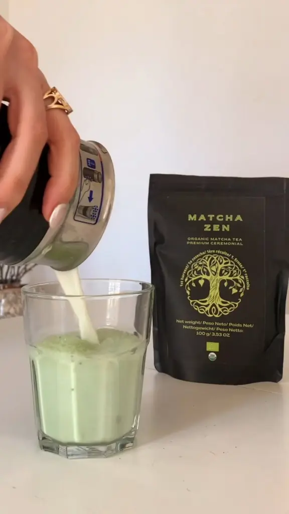 Milk being poured into a glass of iced green matcha beside a black Matcha Zen pouch