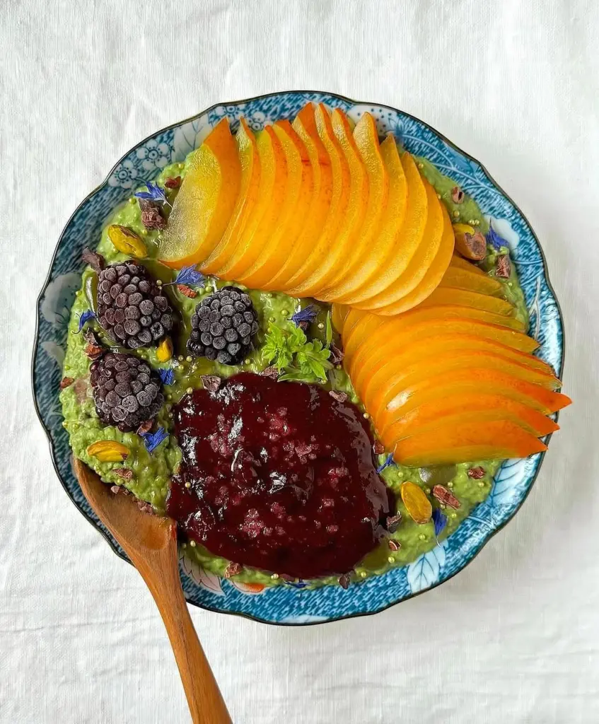 Green bowl topped with sliced orange fruit, frozen blackberries, and red compote