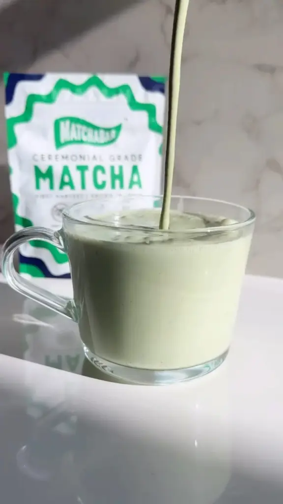 Pale green latte pouring into a clear glass mug with a MatchaBar pouch behind it