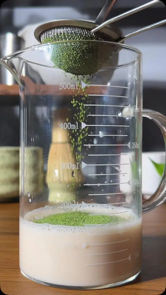 Green powder falling through a metal sieve into a measuring jug of milk