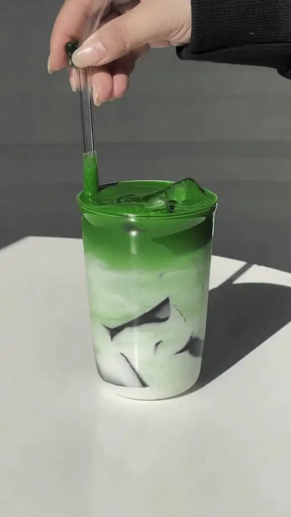 Hand stirring a layered green and white iced drink in a clear glass