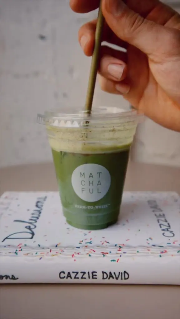 Hand stirring a green drink in a Matchaful cup resting on a book