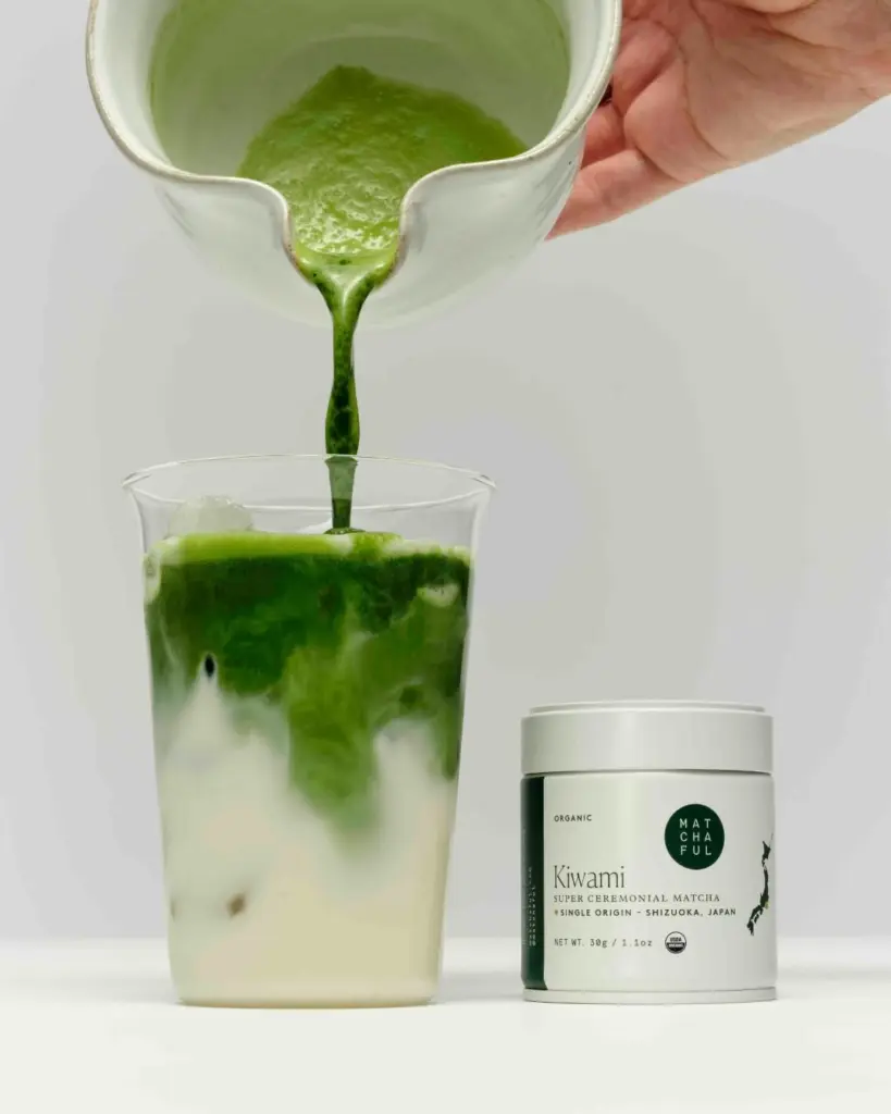 Green matcha poured into a glass of iced milk beside a Kiwami tin