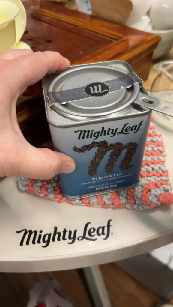 Hand holding a Mighty Leaf Ti Kuan Yin tea tin with a metal infuser resting on top