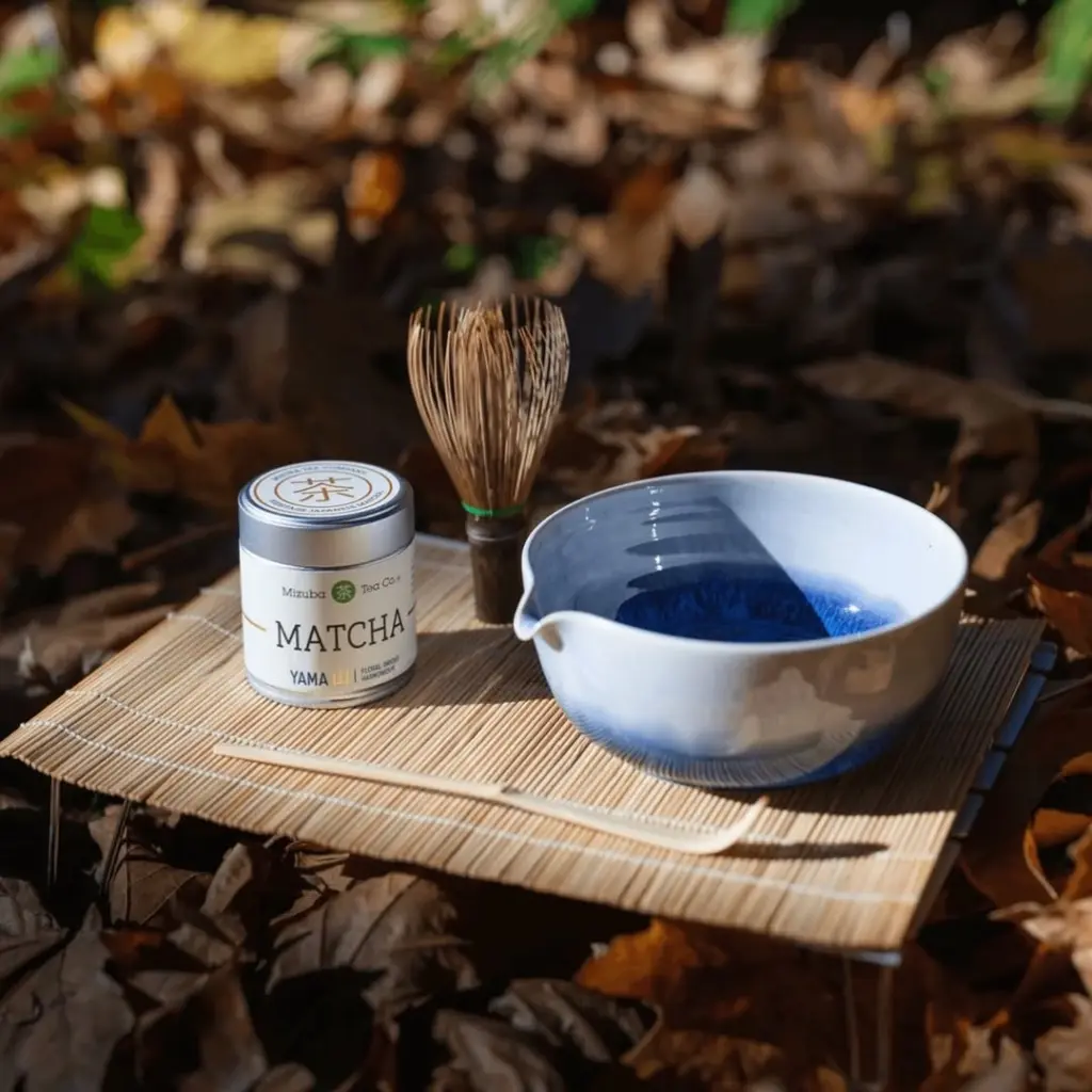 Matcha tin, bamboo whisk, scoop, and blue-and-white bowl among fallen leaves