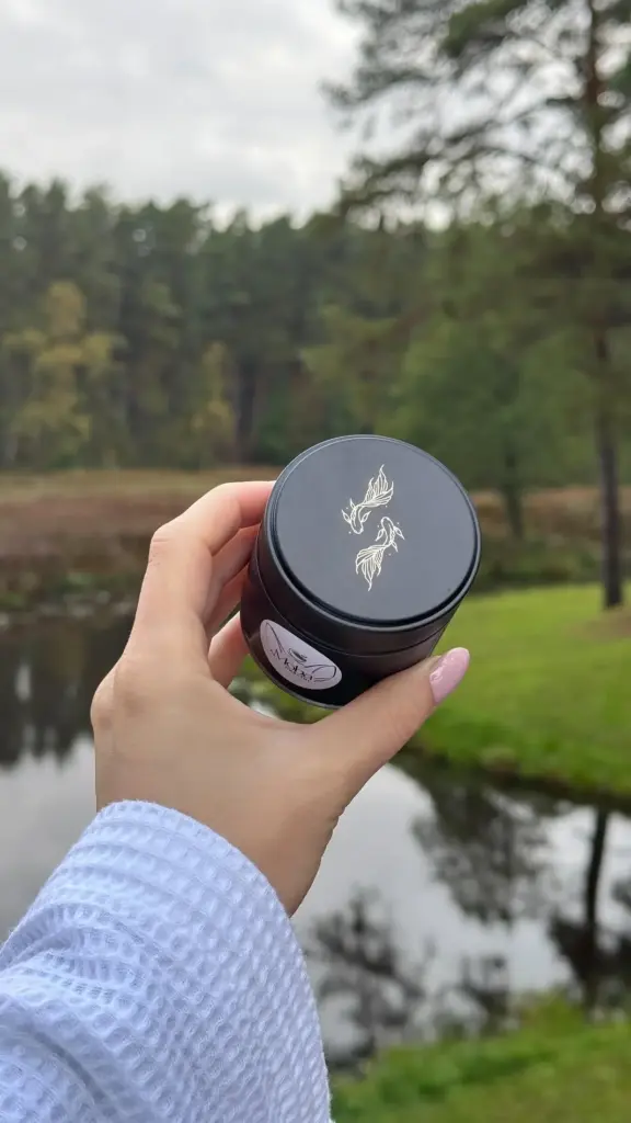 Hand holding a black Moba Matcha tin with a white bird design beside a pond