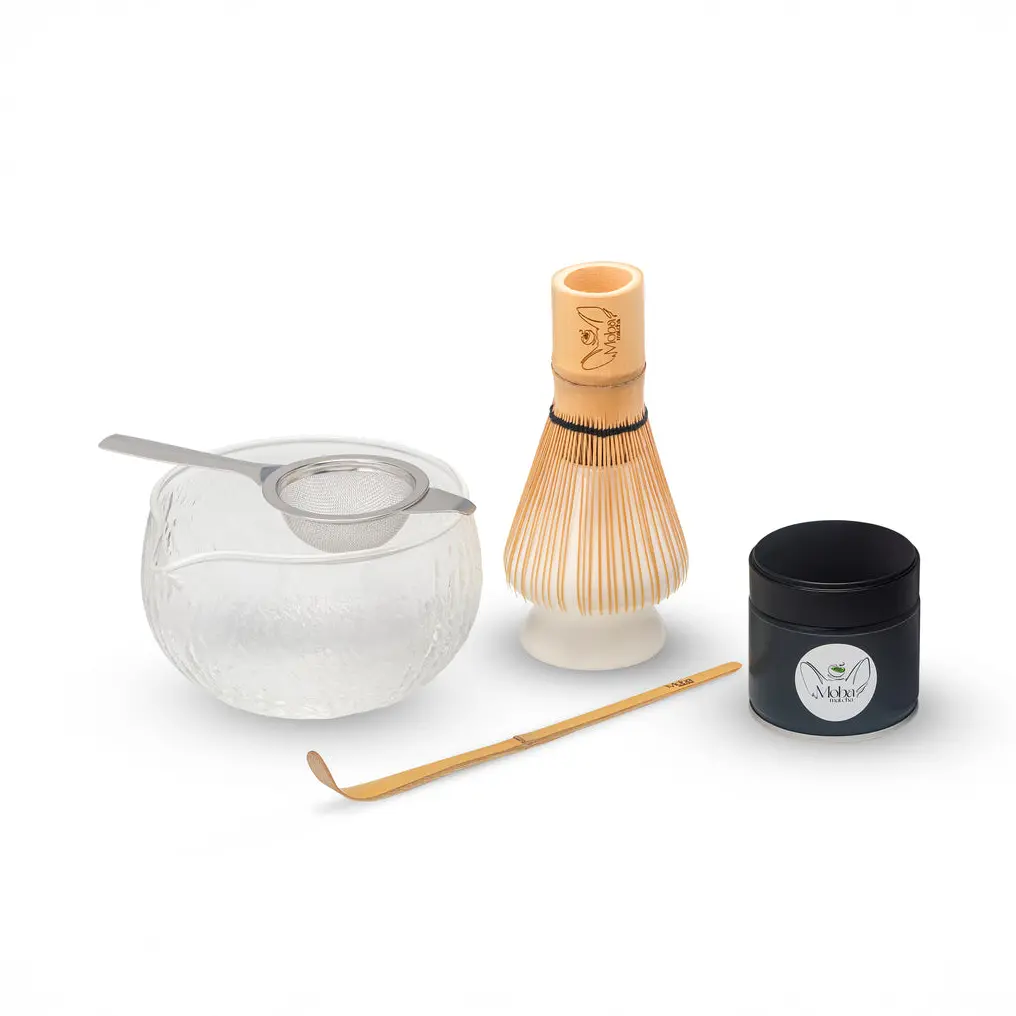 Glass matcha bowl, metal sifter, bamboo whisk on a holder, bamboo scoop, and black Moba Matcha tin
