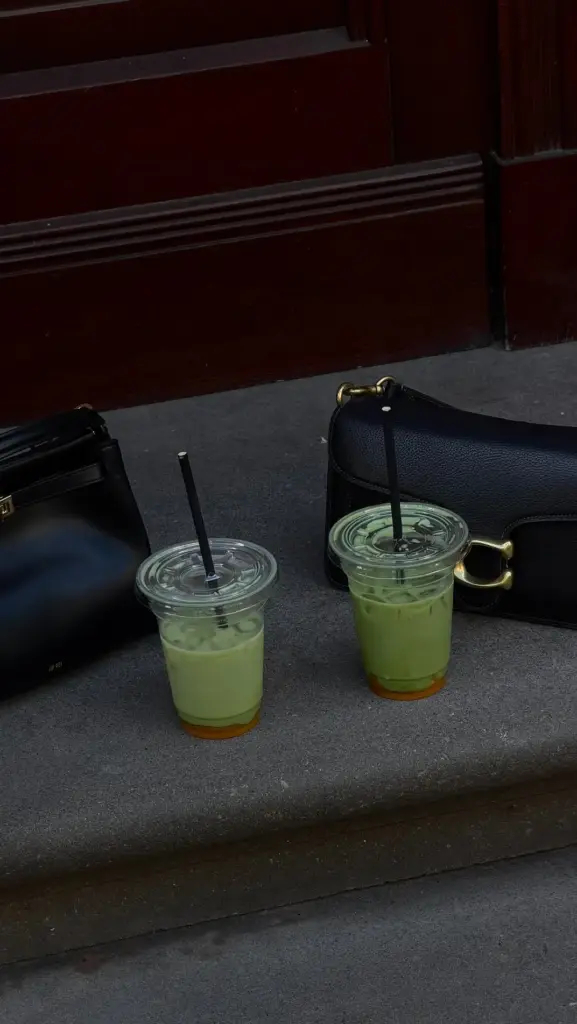 Two iced green matcha drinks with black straws on a stone ledge beside black handbags