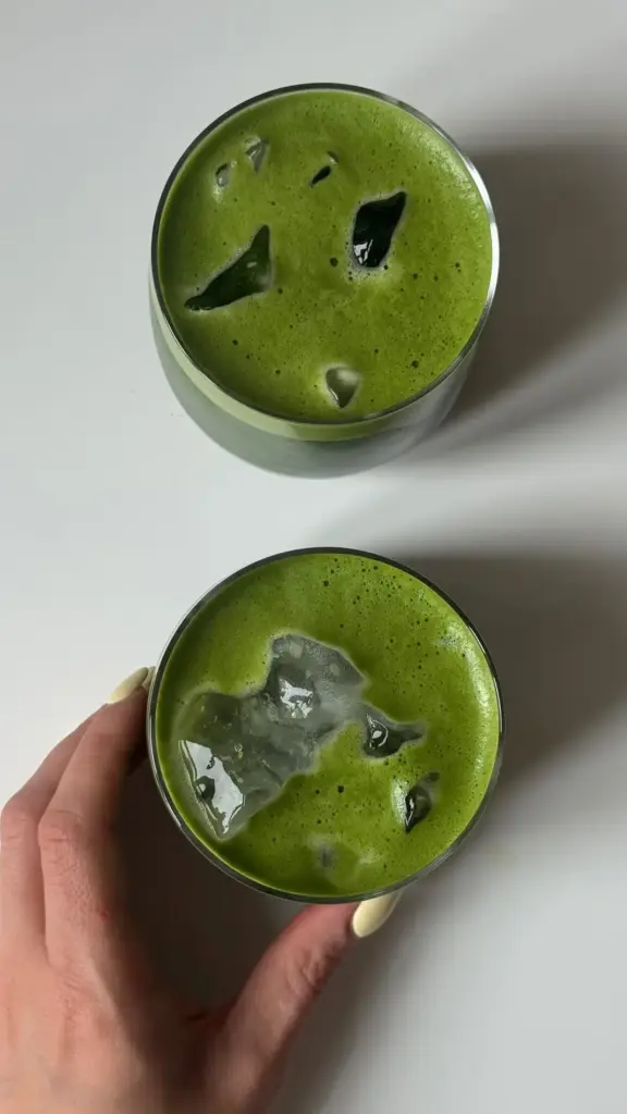Two glasses of frothy green matcha with ice, one held by a hand