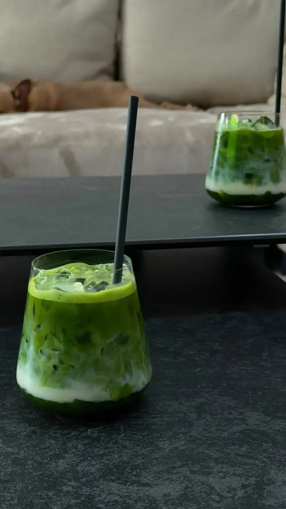 Two glasses of layered green matcha drinks with milk, ice, and black straws on dark tables
