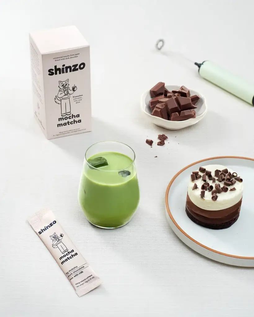 Shinzo Mocha Matcha box and sachet beside an iced green drink, chocolate pieces, and a layered dessert