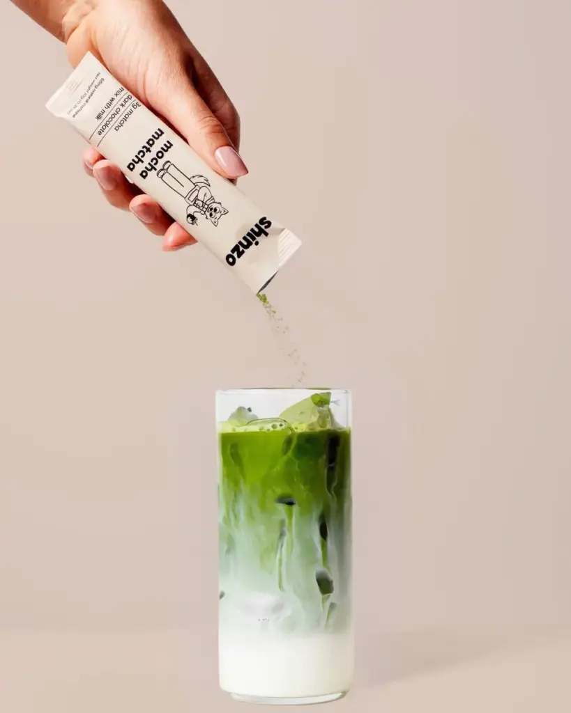 Hand pouring Shinzo Mocha Matcha powder from a single-serve pouch over an iced green drink with milk