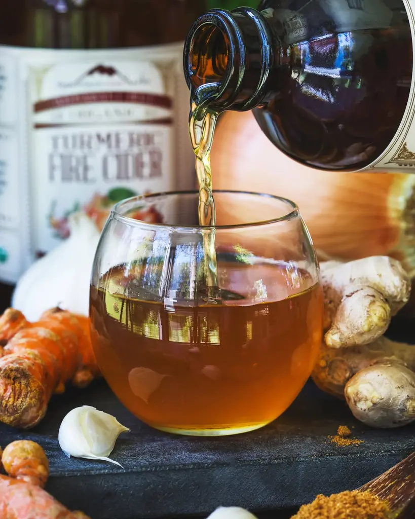 Amber liquid pouring into a glass beside turmeric, ginger, garlic, and ground spice