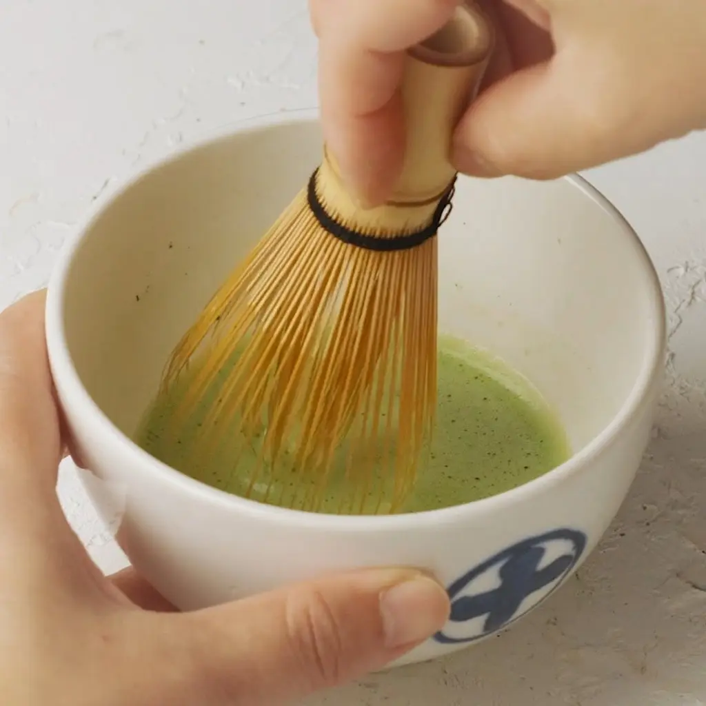 Hands whisking green matcha in a white ceramic bowl