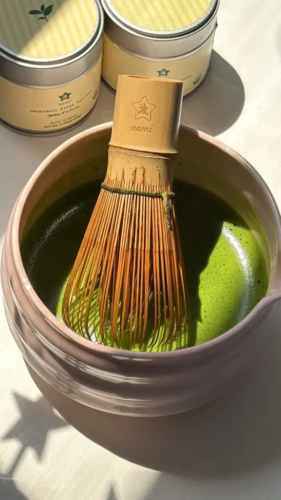 Bamboo whisk resting in prepared matcha with two Nami tins behind it
