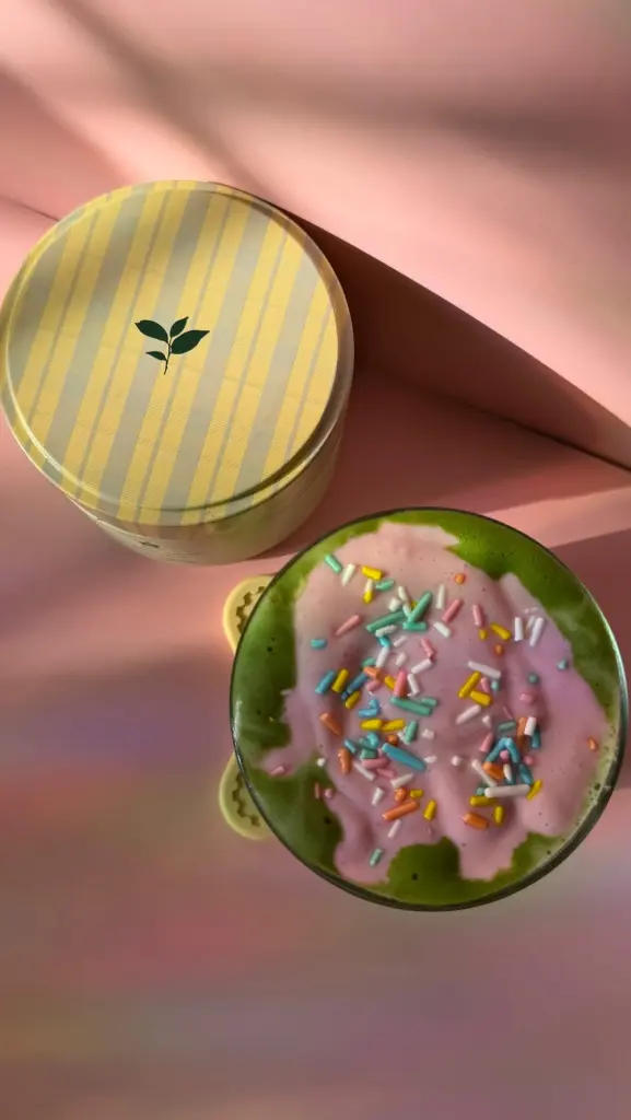 Green drink topped with pink foam and colorful sprinkles beside a striped Nami tin