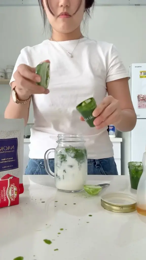 Person adding green matcha mixture to an iced milk drink in a glass mug