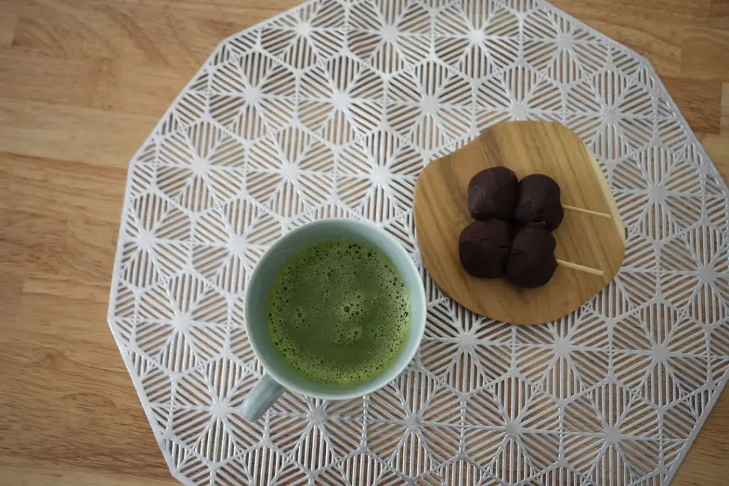 Cup of frothy green tea beside four dark sweets on skewers