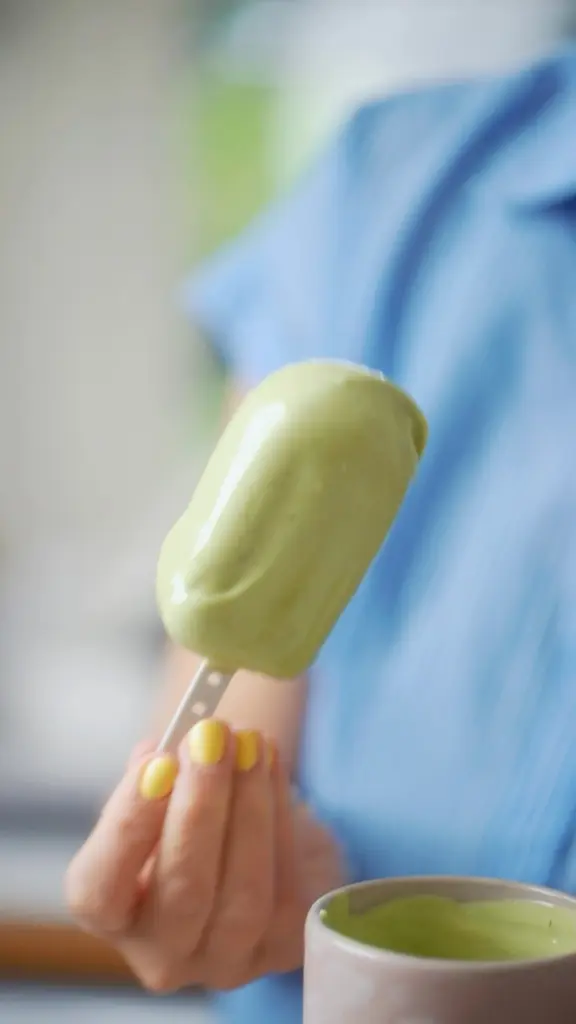 Hand holding a pale green ice pop above a cup of green mixture