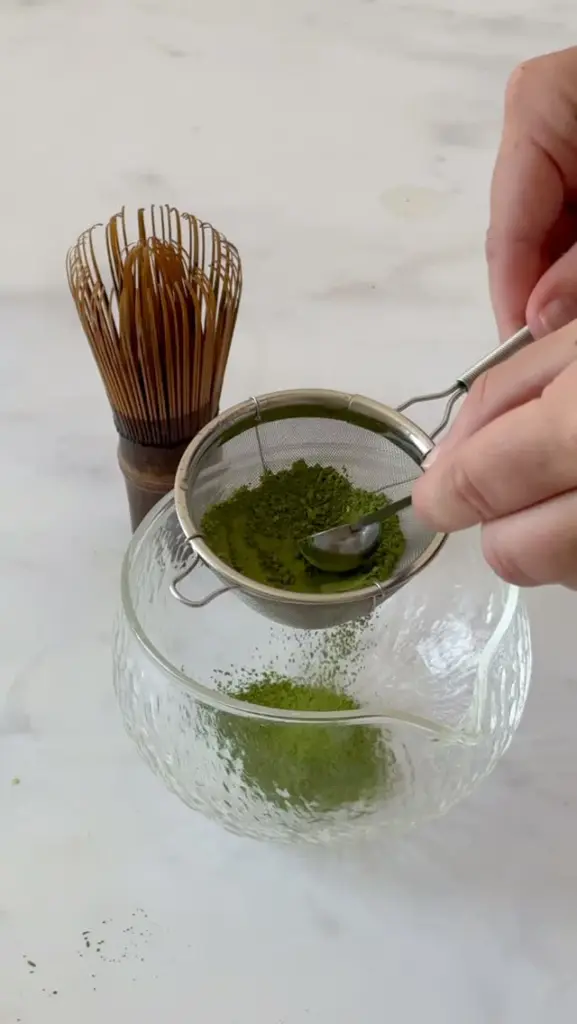Hands pressing green matcha powder through a sieve over a glass bowl