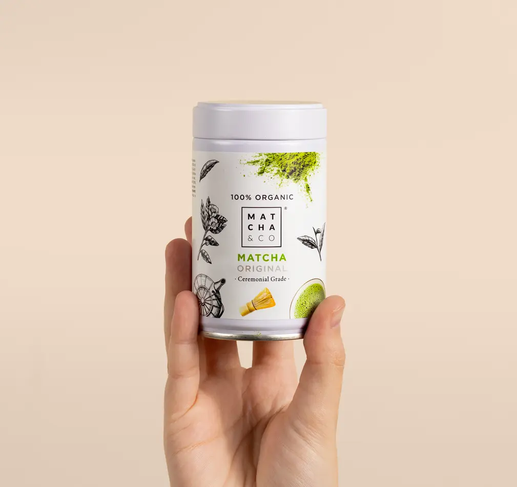 Hand holding a white Matcha & CO Original Matcha Tea canister against a beige background