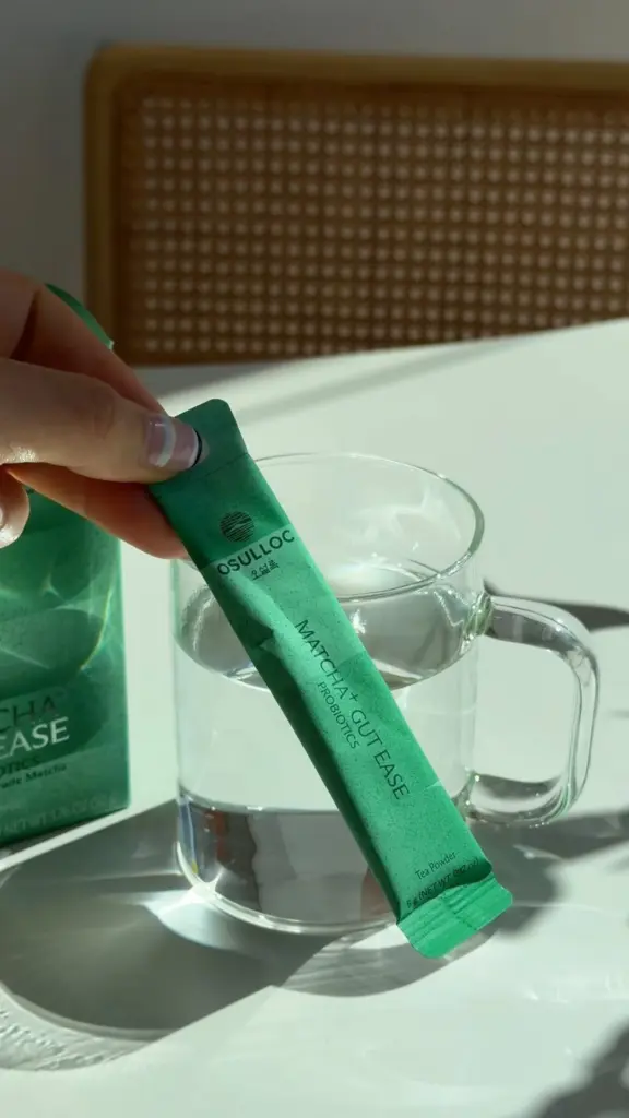 Hand holding an O'sulloc Matcha+ Gut Ease stick over a glass mug of water