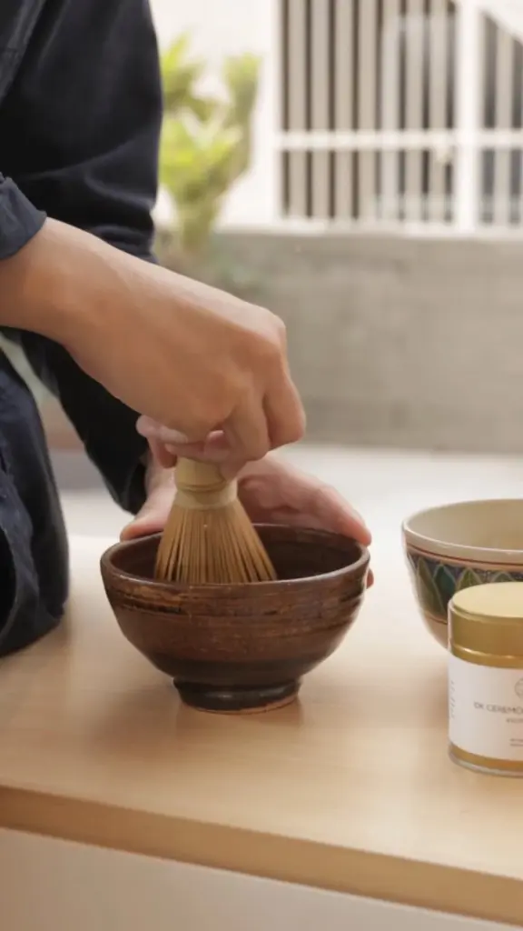 Hands using a bamboo whisk in a brown ceramic tea bowl