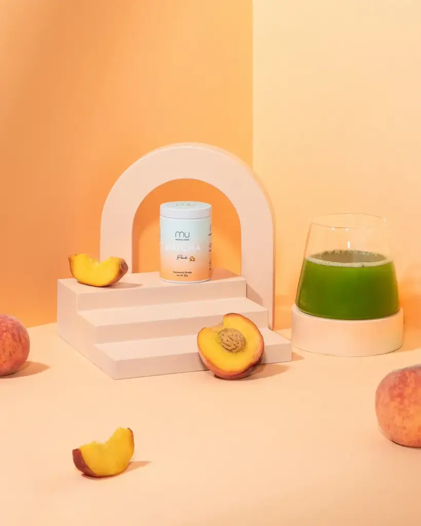 Peach Matcha tin beside a glass of green matcha and sliced peaches on a peach-colored set