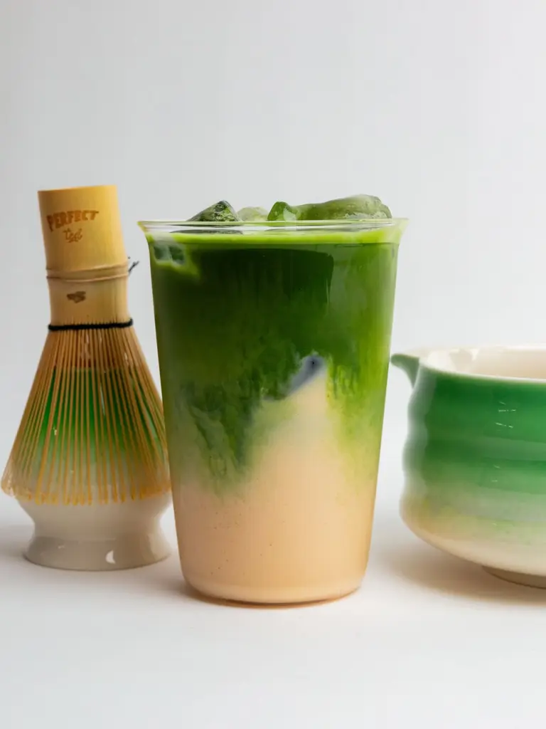 Layered green and cream iced matcha drink beside a bamboo whisk and ceramic bowl