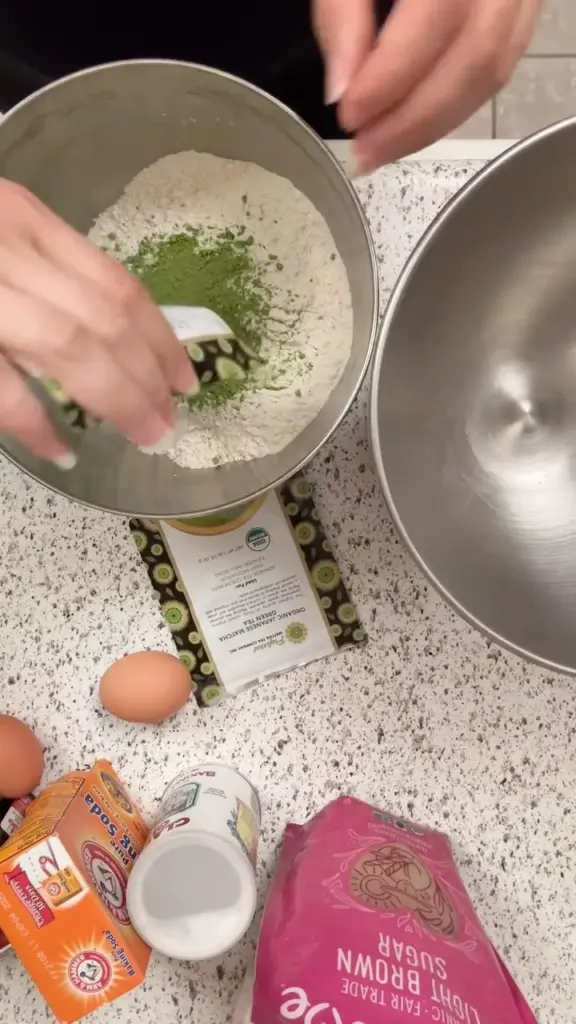Hands adding green matcha powder to a bowl of flour beside eggs and baking supplies