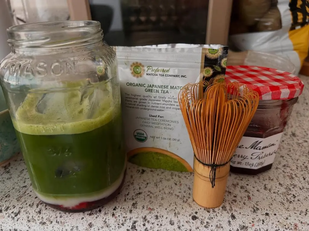 Mason jar with a green drink layered over white and red ingredients beside a matcha pouch and whisk